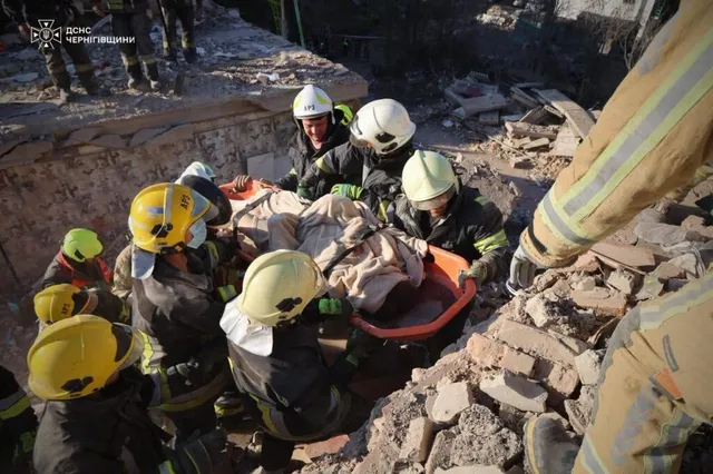 Mai mulți salvatori cu căști de protecție și uniforme de lucru transportă o persoană pe o plăcuță portocalie printre dărâmături de cărămidă.