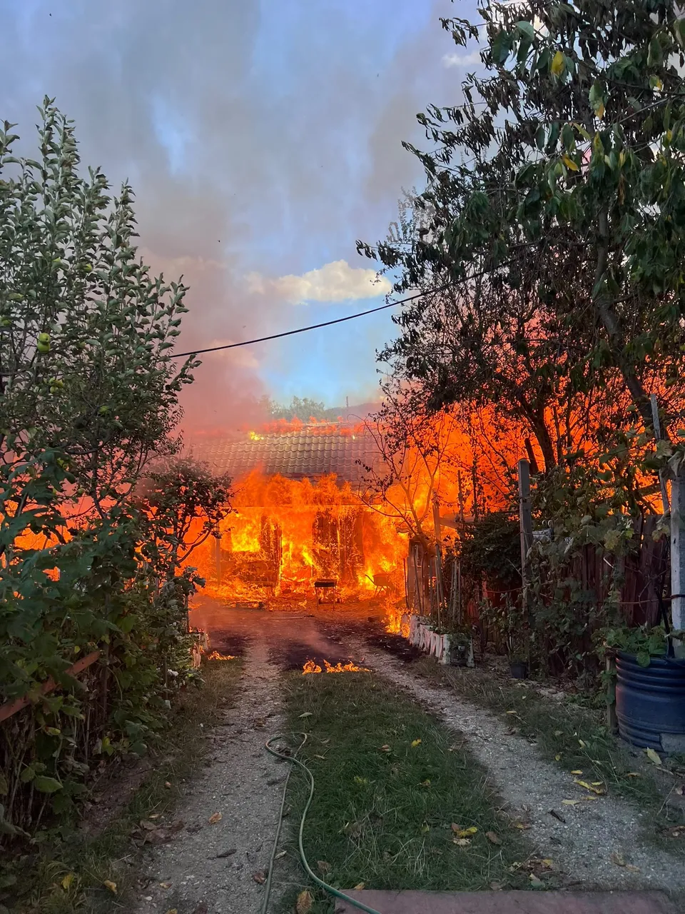 O casă este cuprinsă de flăcări, cu focul vizibil prin ferestre și acoperiș, iar fumul se ridică spre cerul înnorat. În prim-plan, o alee de pământ duce spre clădirea în flăcări, înconjurată de vegetație.