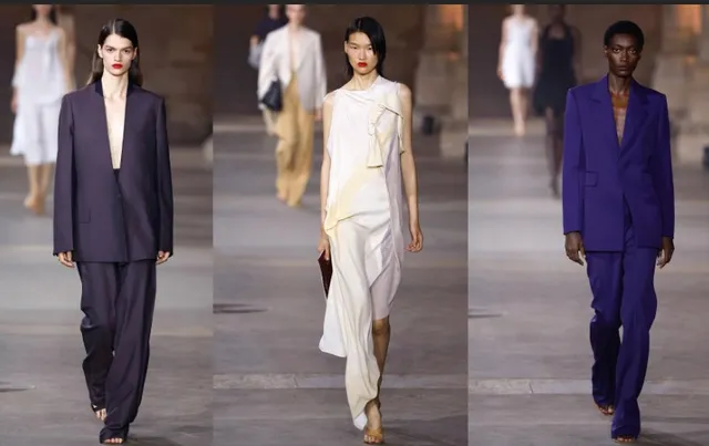 Trei modele de pe un podium poartă haine de modă, inclusiv un costum violet, o rochie albă lungă și un costum închis la culoare.
