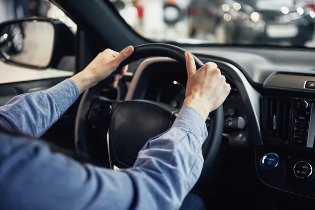 O persoană cu mâinile pe volanul unei mașini, purtând o cămașă albastră, stând în interiorul unui vehicul.