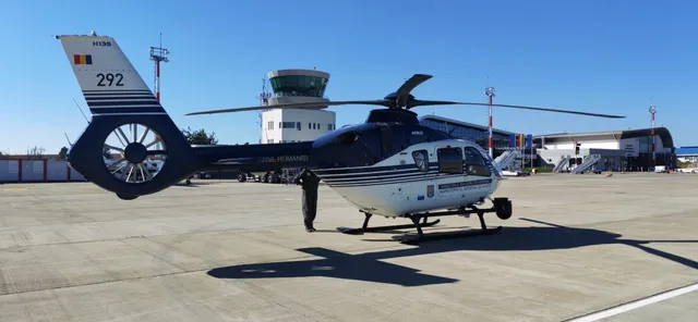 Un elicopter alb și albastru cu numărul 292 pe coadă stă pe o pistă de beton sub un cer senin.