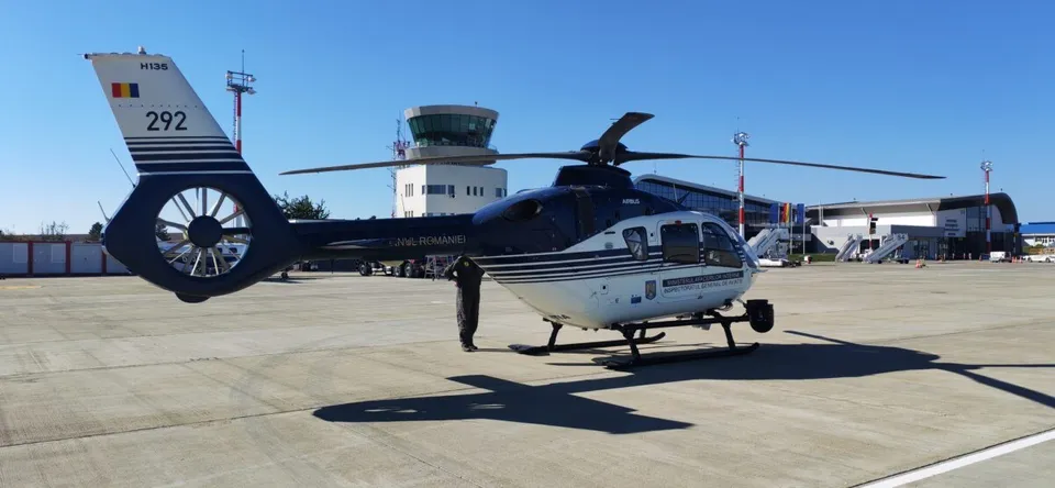 Un elicopter alb și albastru cu numărul 292 pe coadă stă pe o pistă de beton sub un cer senin.