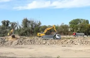 Două excavatoare galbene și două camioane cu bena sunt poziționate pe un teren cu grămezi de pământ și pietriș sub un cer senin.