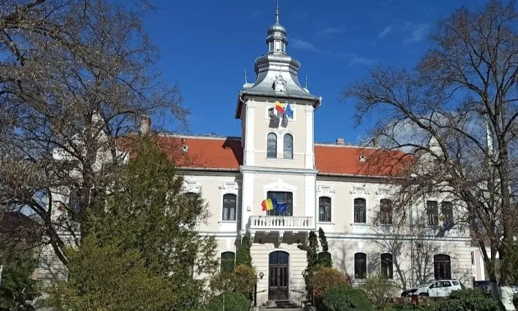 O clădire cu acoperiș roșu și un turn central sub un cer albastru, înconjurată de copaci fără frunze.