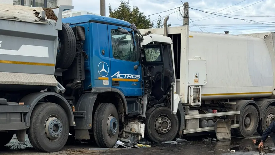 Două vehicule de mare tonaj, un camion albastru și unul alb, sunt implicate într-o coliziune frontală pe un drum asfaltat.