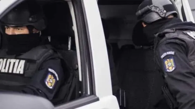 Un polițist cu cască și mască neagră privește spre exterior dintr-o mașină, în timp ce un alt agent în uniformă întunecată stă lângă portiera deschisă.