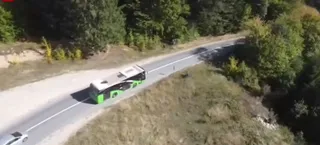 Un autobuz verde și alb circulă pe un drum asfaltat care traversează o zonă cu vegetație și copaci.