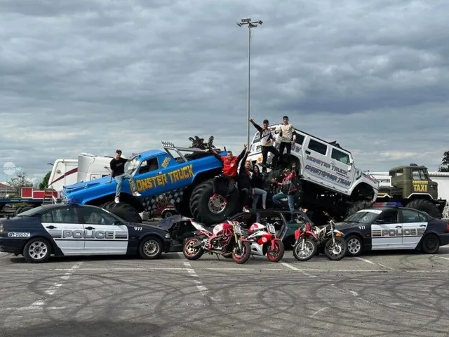 Un monster truck albastru și unul albi cu persoane pe ele stau într-o parcare lângă mașini de poliție și motociclete sub un cer înnorat.