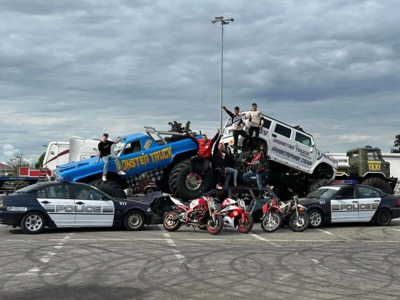 Un monster truck albastru și unul albi cu persoane pe ele stau într-o parcare lângă mașini de poliție și motociclete sub un cer înnorat.