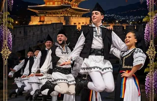 Un grup de dansatori în costume tradiționale albe și negre execută un dans în aer în fața unei clădiri iluminat noaptea.