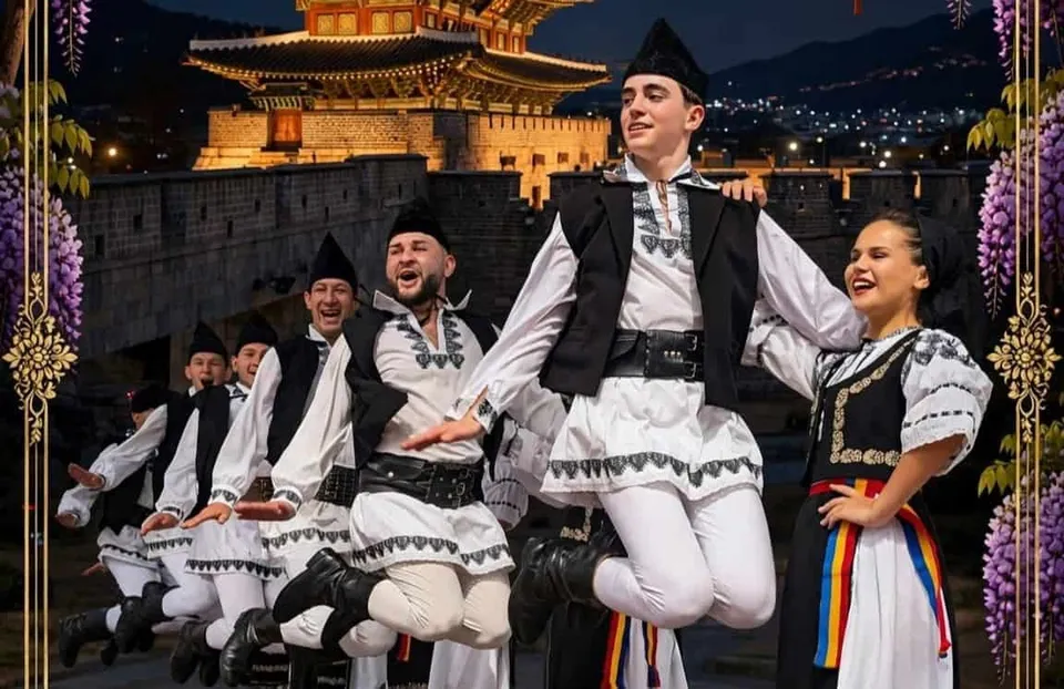 Un grup de dansatori în costume tradiționale albe și negre execută un dans în aer în fața unei clădiri iluminat noaptea.