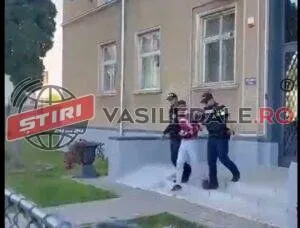 Două persoane în haine închise și o persoană cu tricou roșu merg pe o scară în fața unei clădiri cu ferestre albe.