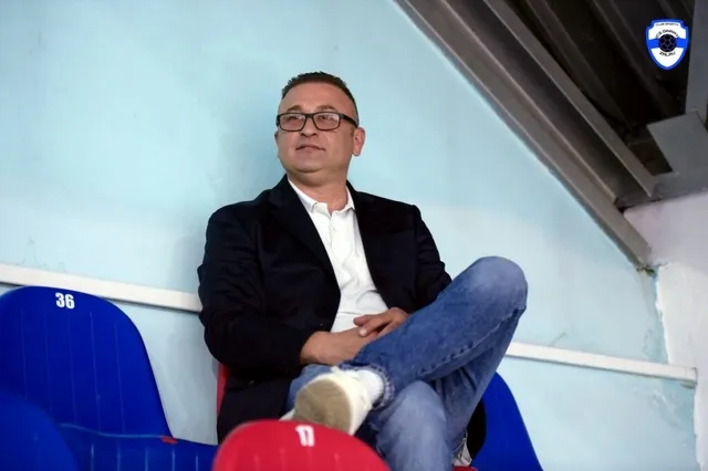 Un bărbat cu ochelari, purtând sacou negru, cămașă albă și blugi, stă așezat pe un scaun de tribune albastru.