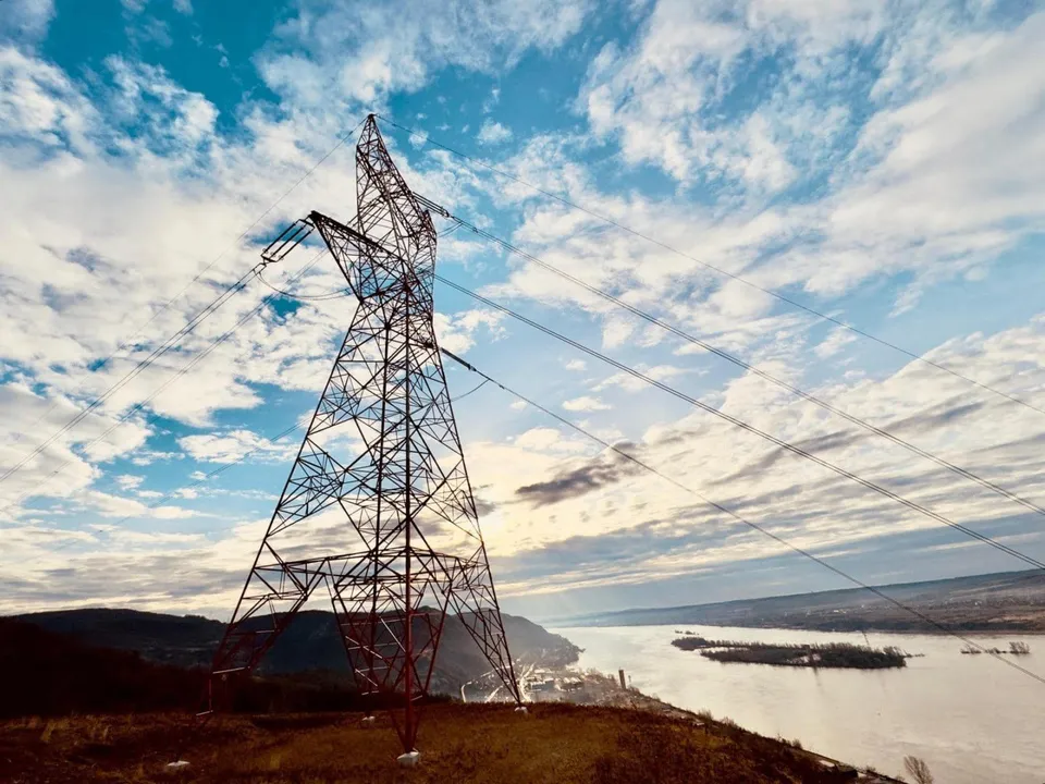 O turnul de înaltă tensiune metalic se ridică pe un deal lângă un fluviu sub un cer cu nori.
