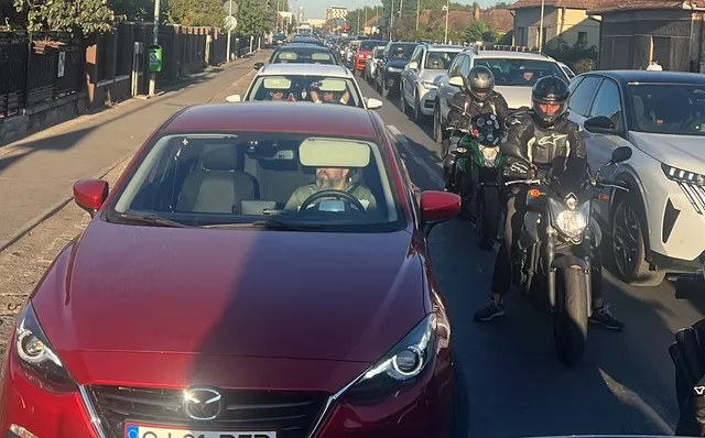 O linie de mașini și motociclete circulă pe un drum asfaltat lângă un trotuar și garduri de proprietăți.