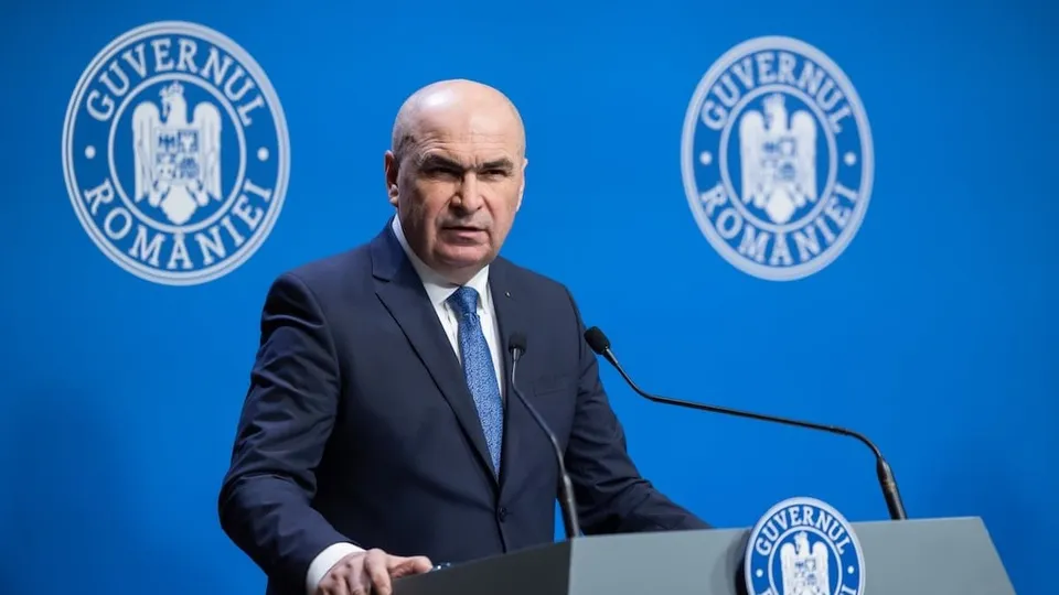 Un bărbat cu părul scurt și costum închis la culoare stă în spatele unui podium în fața unui fundal albastru cu sigilii oficiale.