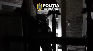 Silueta unui agent de poliție în uniformă stă în fața unei uși deschise într-un decor nocturn.