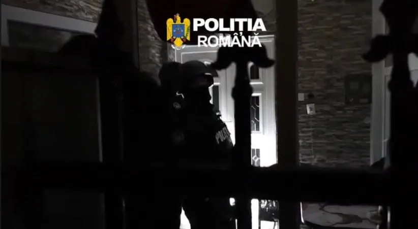 Silueta unui agent de poliție în uniformă stă în fața unei uși deschise într-un decor nocturn.