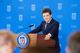 Un bărbat într-un costum albastru și cămașă albă stă în spatele unui podium de lemn în fața unui fundal albastru cu embleme.