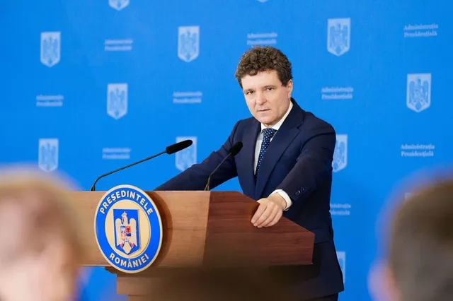 Un bărbat într-un costum albastru și cămașă albă stă în spatele unui podium de lemn în fața unui fundal albastru cu embleme.
