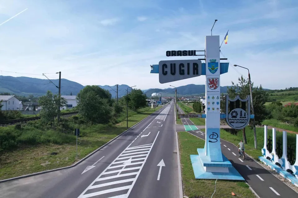 O stradă asfaltată cu marcaje albe traversează un peisaj verde sub un cer senin, având un monument vertical albastru și alb lângă drum.