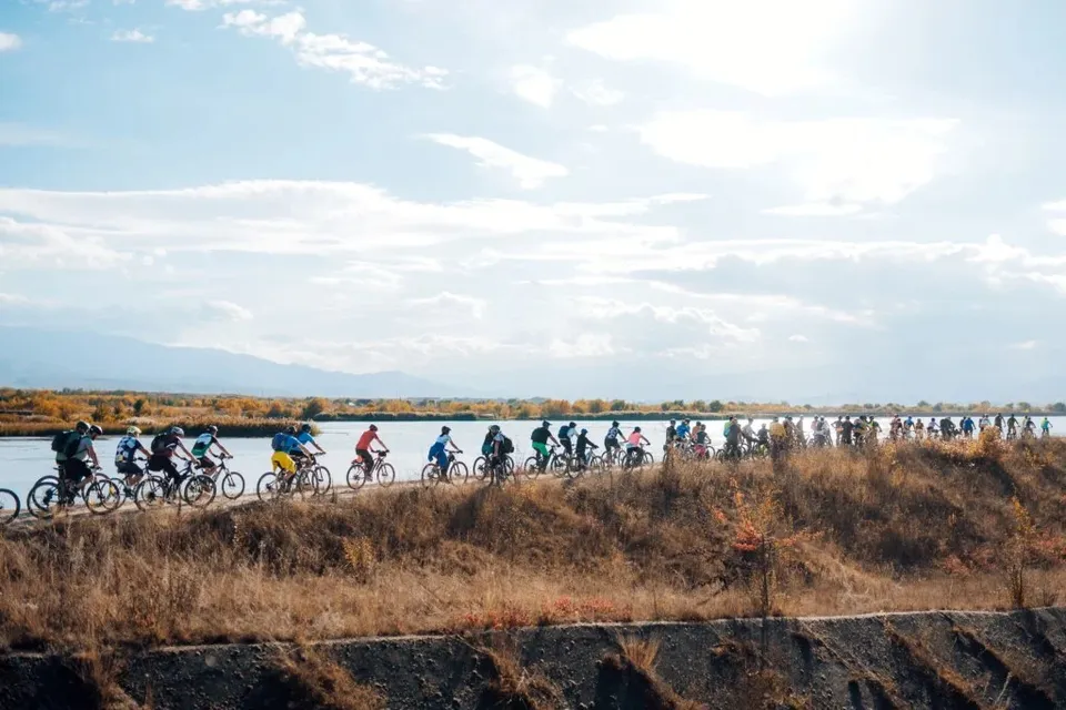 O mulțime de bicicliști în echipamente colorate pedalează pe un drum de pământ de lângă un lac sub un cer cu nori.
