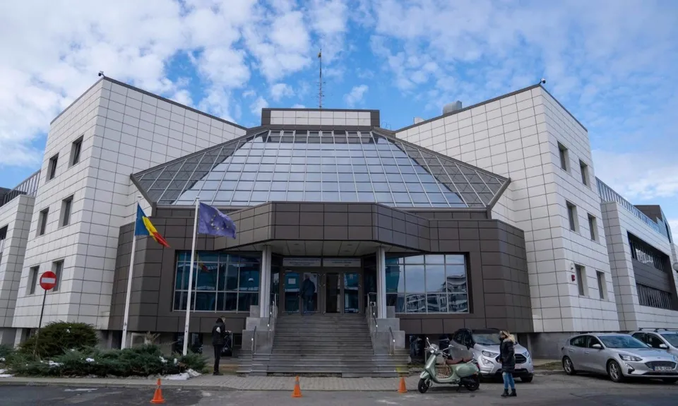 O clădire modernă cu fațadă albă și gri, cu o structură de sticlă în centru, sub un cer cu nori.