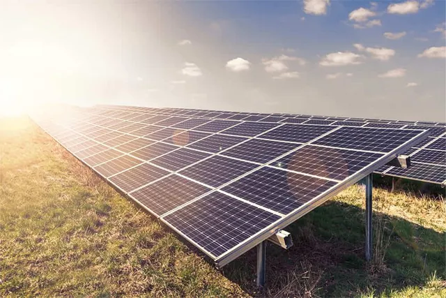 Rânduri de panouri solare sunt instalate pe un câmp de iarbă sub un cer cu nori și lumină solară puternică.