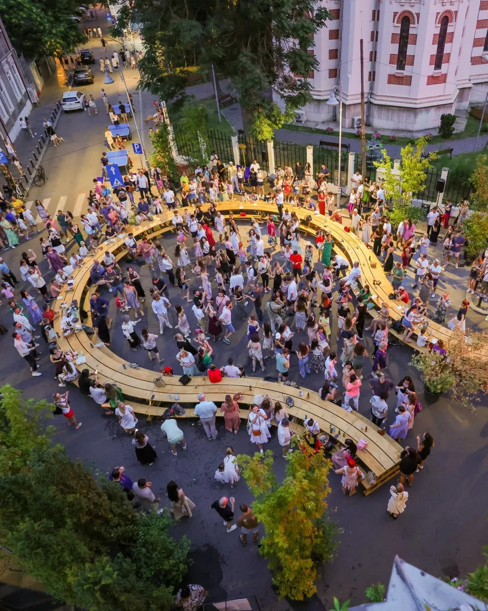 O mulțime de oameni stă la mese lungi de lemn dispuse sub formă de cerc pe o stradă urbană, înconjurată de clădiri și copaci.