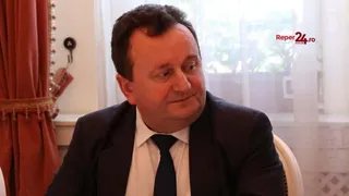 Un bărbat în costum și cravată este înfățișat în plan apropiat, privind spre partea dreaptă, într-o cameră cu draperii și un geam din spate.