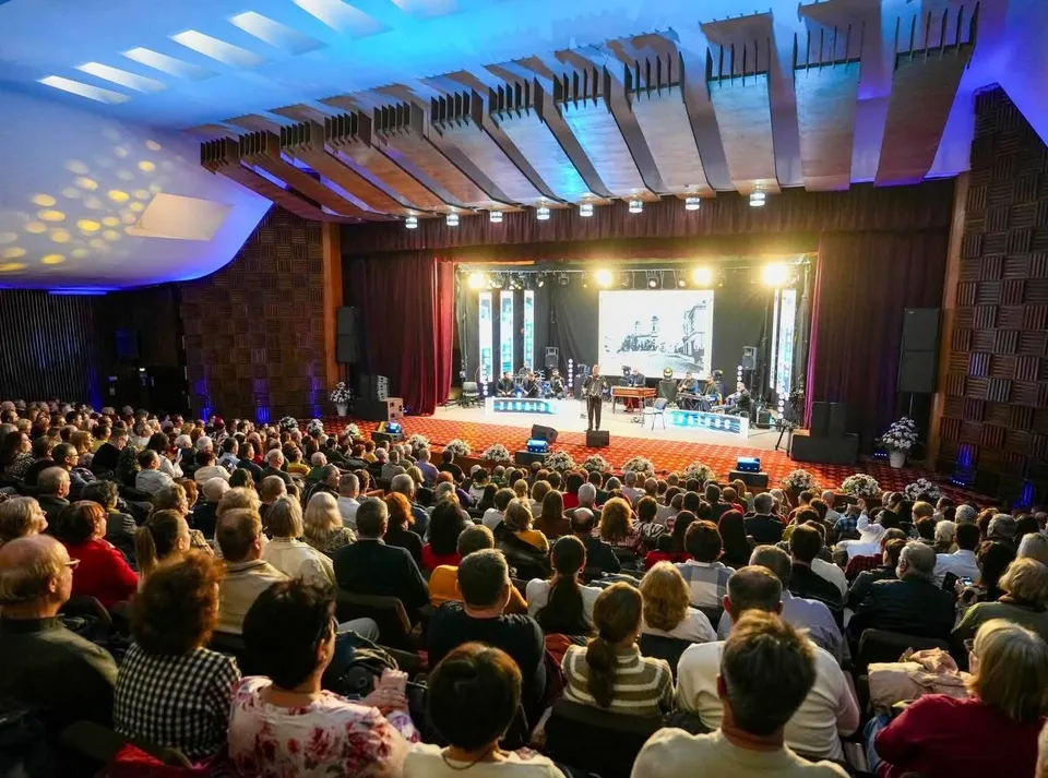 O mulțime de oameni stă așezată în sală, privind spre o scenă iluminată unde se află un instrument muzical și un ecran mare.