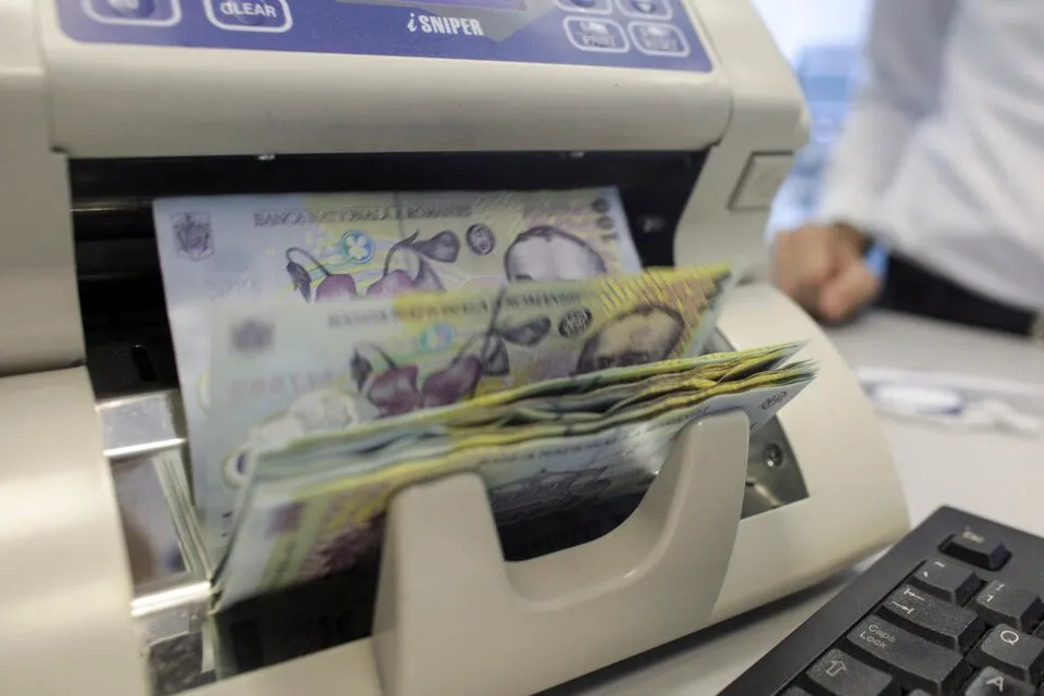 Banii de tip bancnotă sunt introduși într-un aparat de numărat bani de culoare albă, lângă un tastatură neagră.