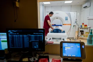 Un medic în halat roșu stă lângă un pacient întins pe o masă de scanare într-o cameră medicală cu un aparat CT și monitoare.