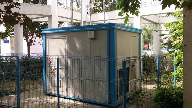 Un container gri cu margini albastre este situat în spatele unui gard metalic albastru, lângă vegetație și sub o structură de beton.