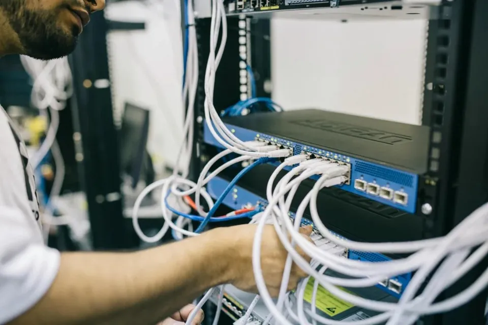 Un bărbat cu tricou alb manipulează cabluri de rețea albe conectate la un echipament de server într-un rack metalic negru.