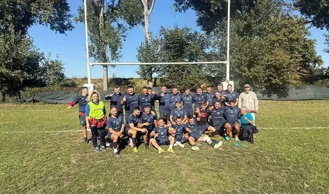 Un grup de sportivi în echipamente de rugby posedă pentru o fotografie pe un teren de iarbă, în fața unui stâlp de marcaj alb.