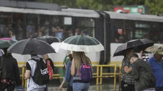 Oamenii merg pe stradă sub umbrele de diverse culori în timp ce plouă, având un tramvai în fundal.
