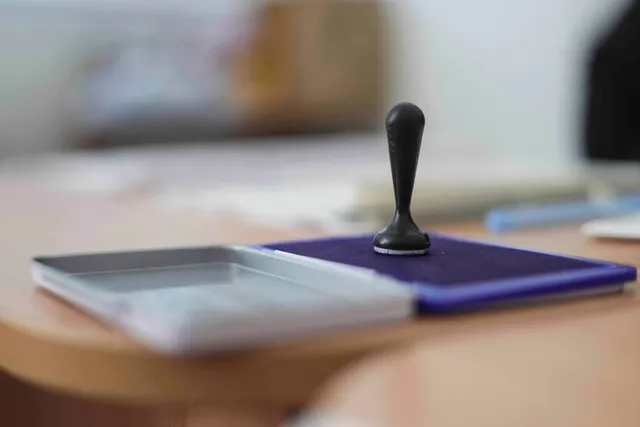 Un ștampilă neagră stă pe o almohadă de cerneală albastră aflată pe o masă de lemn lângă o casetă de ștampilare transparentă.
