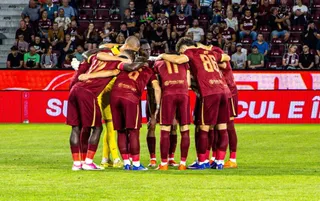 Un grup de fotbaliști în echipamente de culoare roșie și galbenă stau îmbrățișați într-un cerc pe un teren de fotbal.