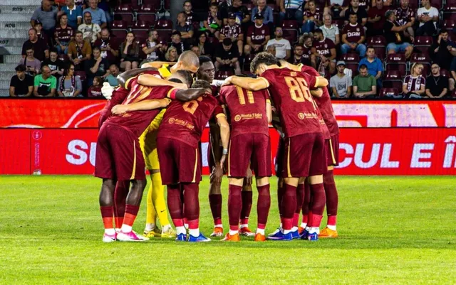 Un grup de fotbaliști în echipamente de culoare roșie și galbenă stau îmbrățișați într-un cerc pe un teren de fotbal.