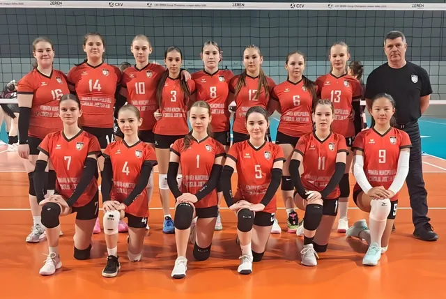 O echipă de volei formată din femei poartă echipamente roșii și negre, așezate în două rânduri pe un teren de volei portocaliu.