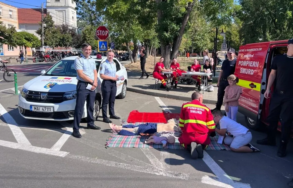 Un grup de persoane în uniforme roșii și albastre execută manevre de prim ajutor pe un om întins pe un covoraș pe asfaltul unei străzi.