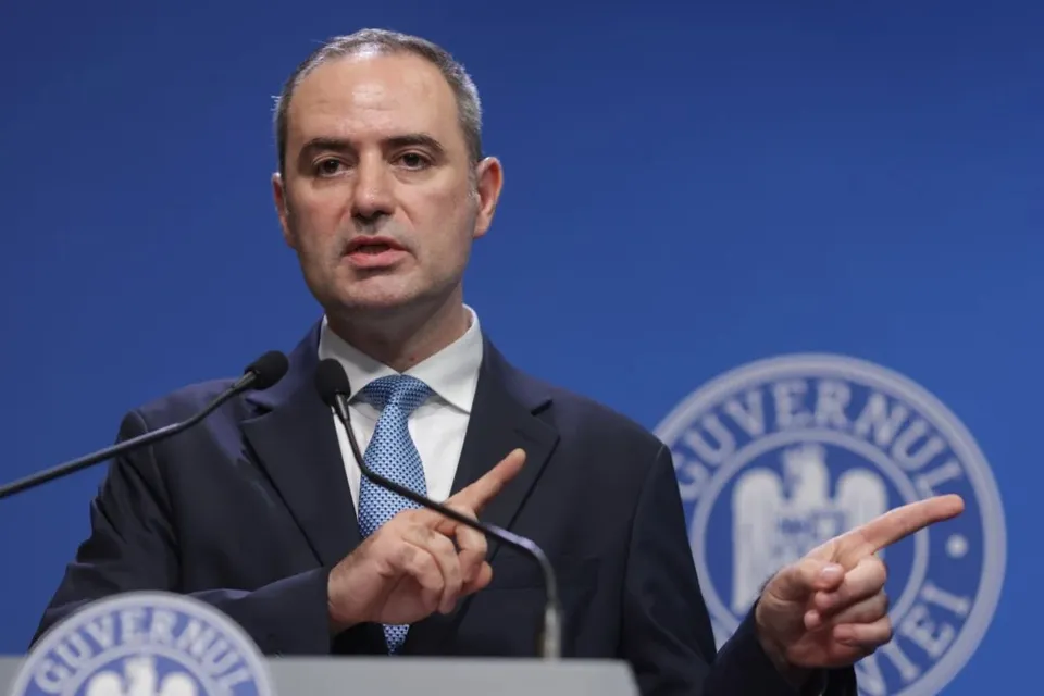Un bărbat cu sacou închis și cămașă albă cu cravată albastră vorbește în fața unui fundal albastru cu un sigiliu circular.