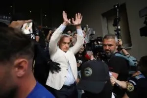 Un bărbat cu sacou alb stă cu mâinile ridicate în timp ce este înconjurat de persoane și camere de filmare.