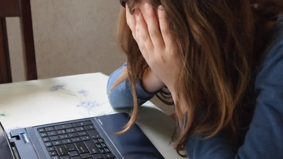 O persoană cu părul lung stă la o masă cu un laptop deschis, având fața acoperită de mâini.