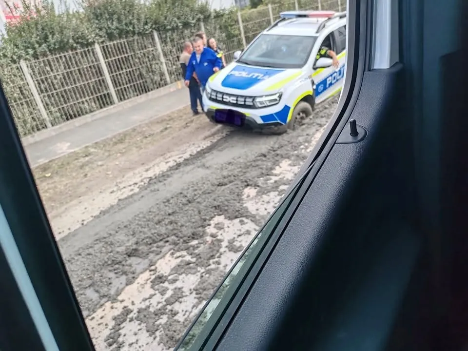 O mașină de poliție albastră și albă este parcată pe o suprafață de beton proaspăt turnat lângă un gard metalic și câțiva oameni stau în apropiere.