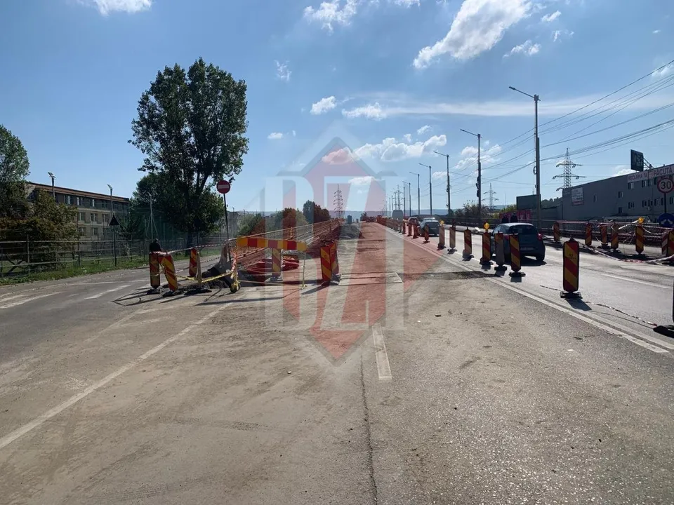 O porțiune de drum este delimitată de bariere portocalii și conuri de circulație lângă o zonă de șantier sub un cer înnorat.