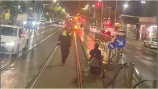 O persoană aflată într-un scaun cu rotile și un agent de poliție se află pe un trotuar lângă șinele de tramvai noaptea.