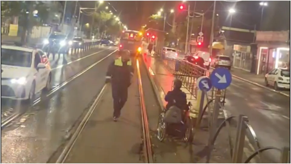 O persoană aflată într-un scaun cu rotile și un agent de poliție se află pe un trotuar lângă șinele de tramvai noaptea.