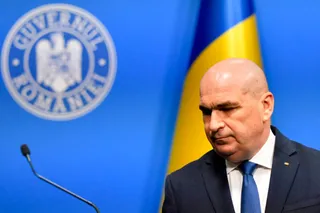 Un bărbat cu părul rare și costum închis la culoare stă în fața unui fundal albastru cu un steag și un sigiliu.